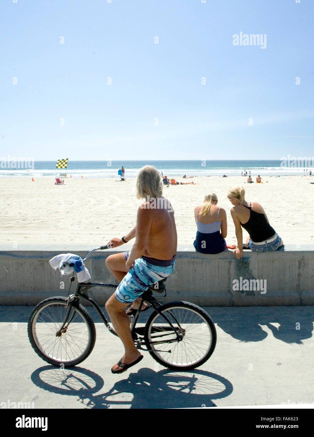 Man cycling along a beach, California with two girls sitting on a wall. Stock Photo