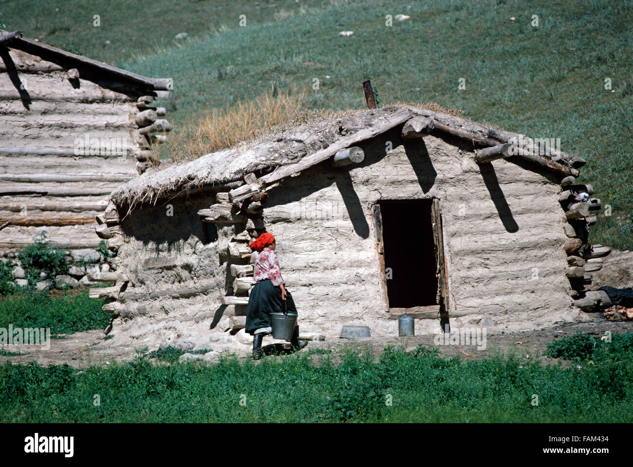 Kazakh woman carrying water buckets from farmhouse built out of wood and mud in hills North of Urumqi, Xinjiang Province, China Stock Photo