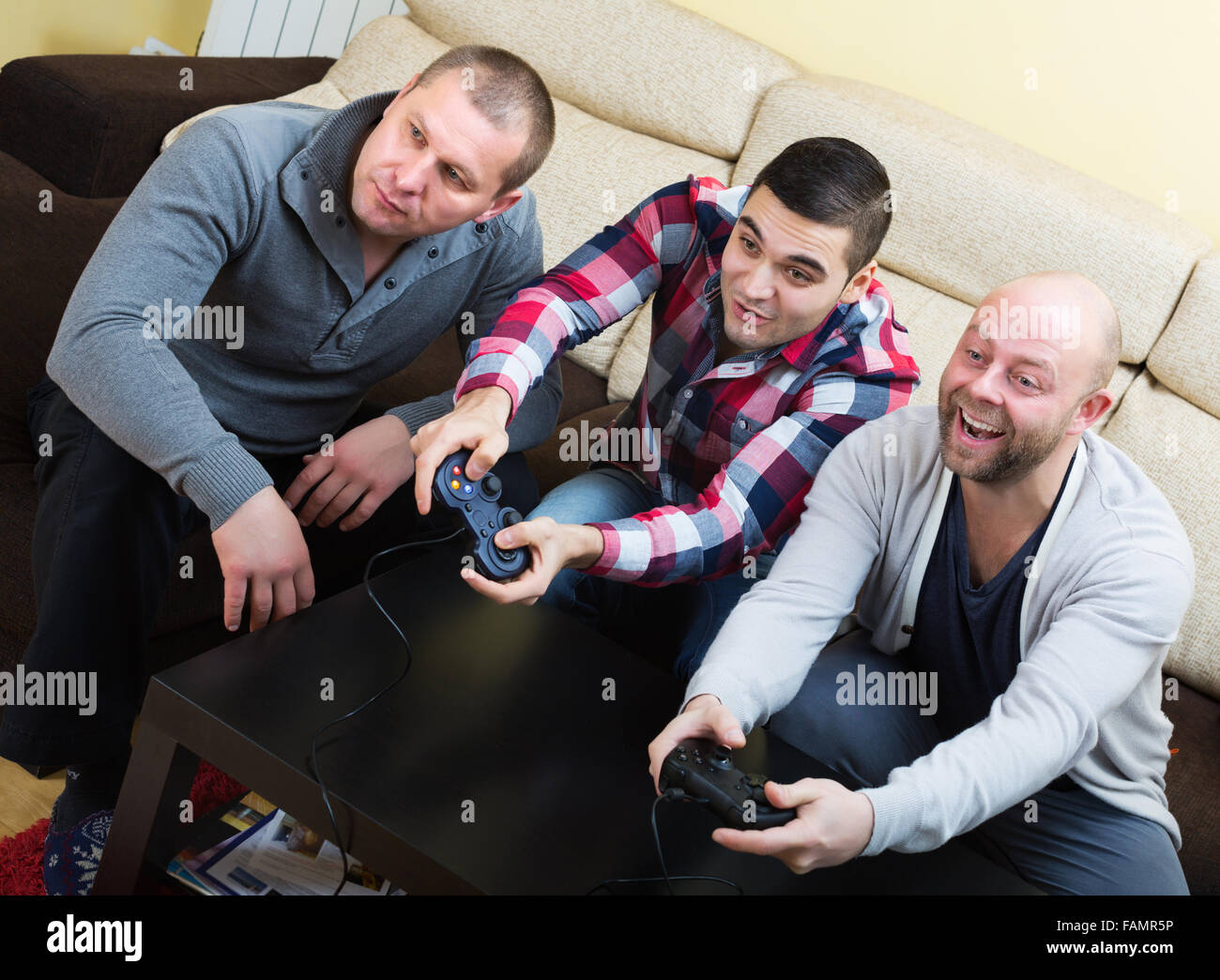 Three heated guys sitting on sofa with joysticks indoors Stock Photo
