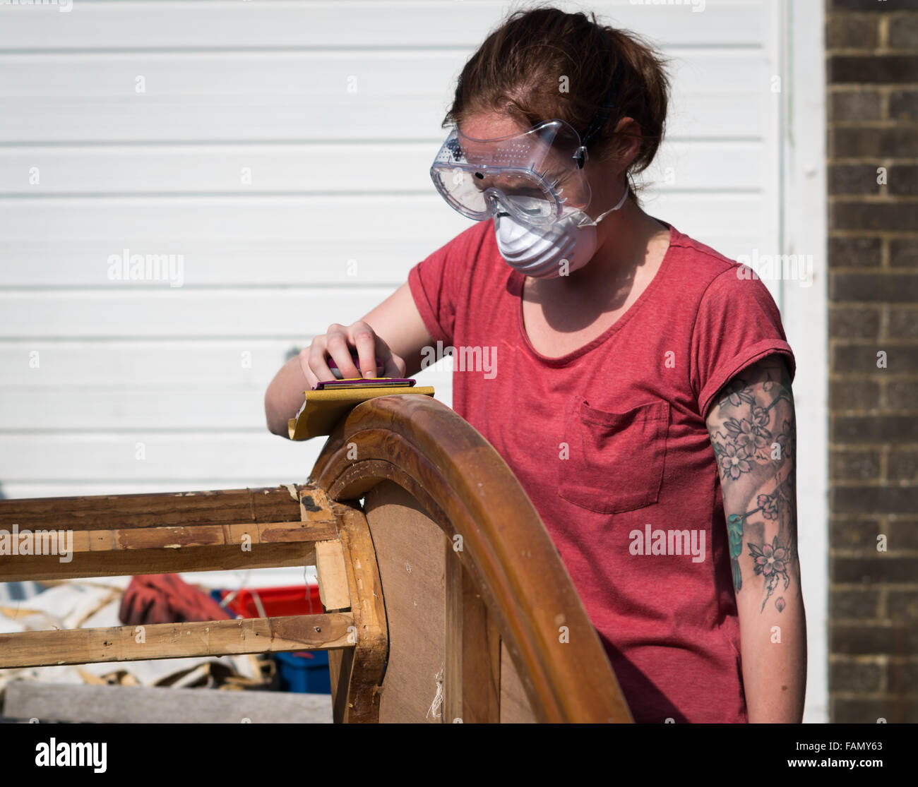 Tattooed young woman sanding by hand a piece of antique wooden furniture outside wearing protective equipment. Stock Photo