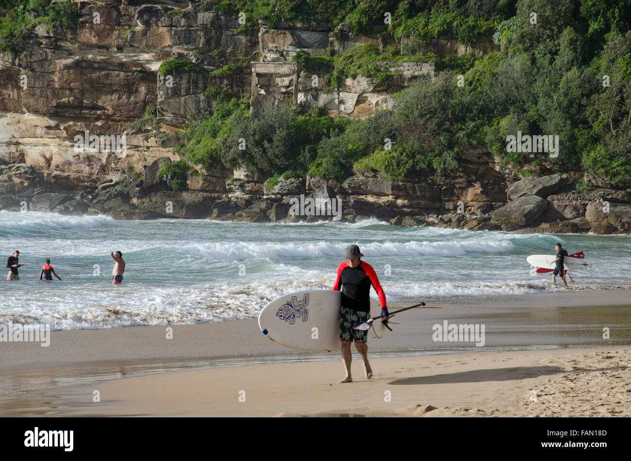 An older man carrying a standup paddleboard (SUP) along Freshwater Beach on a sunny Sydney morning in Australia Stock Photo