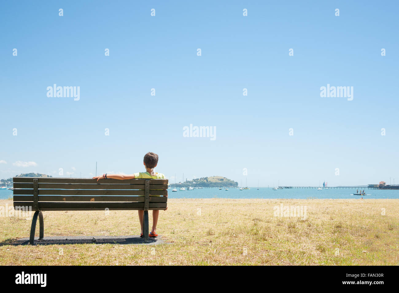 Boy sits on bench seat in the bay. Summer day view across bay, Orakei Auckland, New Zealand under blue sky. Stock Photo