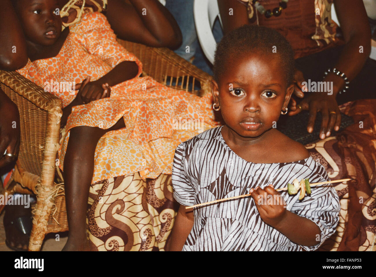A little girl from Gambia holding wooden barbecue stick with vegetables on it. The girl is wearing blue pattern dress. Stock Photo