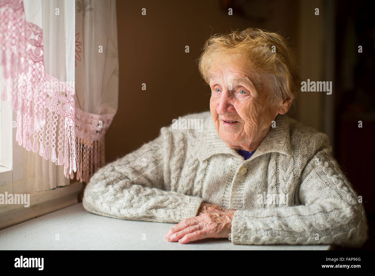 An elderly woman sits at a table in the kitchen near the window Stock