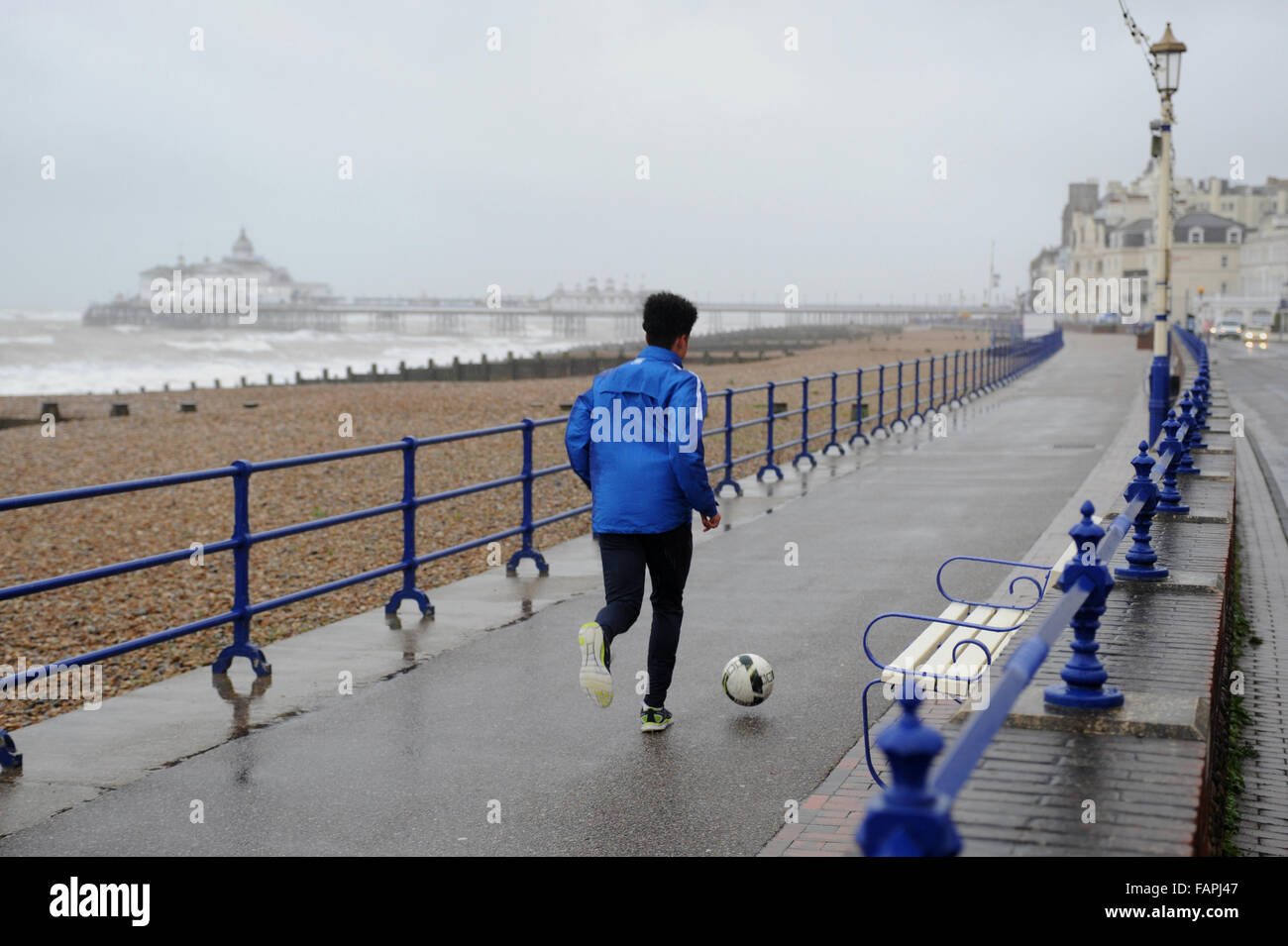A young man kicks a football along the promenade on a wet and windy day