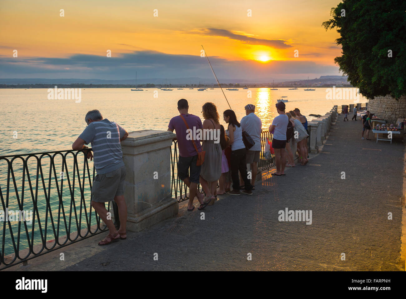 People sunset, view on a summer evening of people gathered on the esplanade in the harbour at Syracuse (Siracusa) to watch a colourful sunset, Sicily. Stock Photo