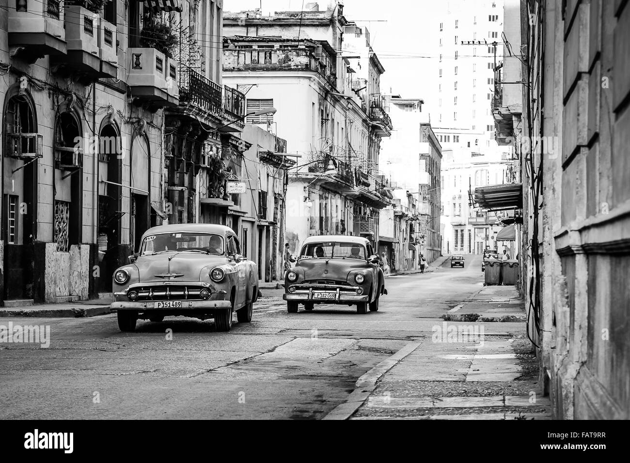 A monochrome image of a duo of classical cars beginning to fill the frame along a typically rugged street in Havana. Stock Photo