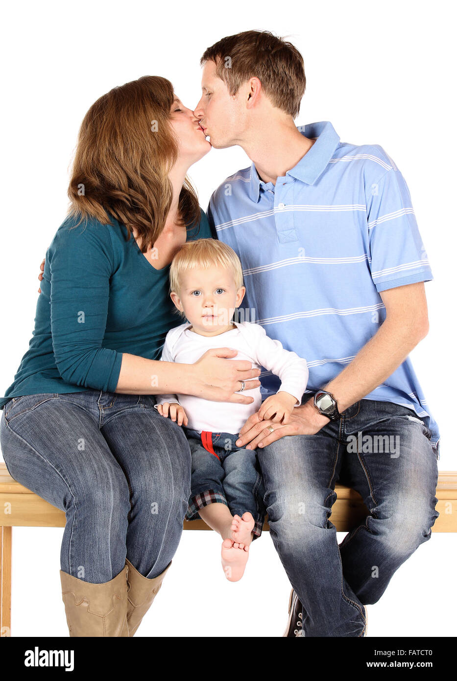 Caucasian family with mom dad and baby boy. They are all wearing denim jeans and sitting on a wooden bench. The parents are kiss Stock Photo