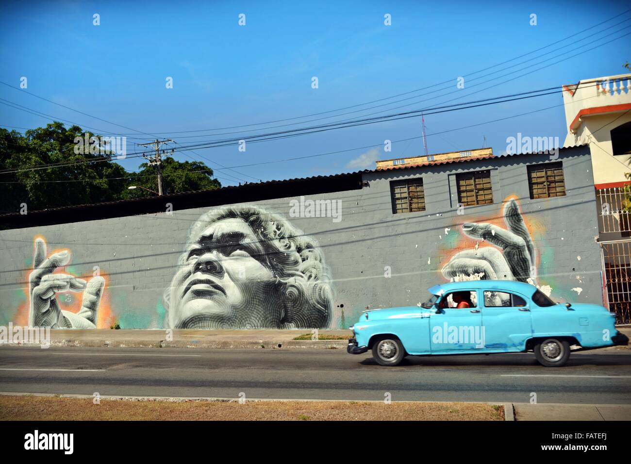 Cuban street art in Havana; mural of an illuminated Cuban womens face and hands upturned to the sky and blue vintage car passing Stock Photo