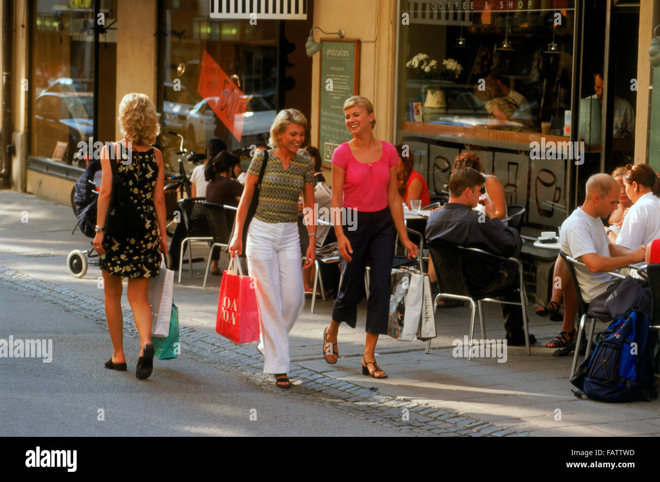 Three Swedish women with shopping bags walking past sidewalk restaurant on Stockholm street in summer Stock Photo