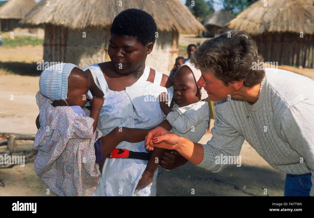 African woman holding two babies in her arms in front of village hut in rural Zimbabwe  visiting with female tourist Stock Photo