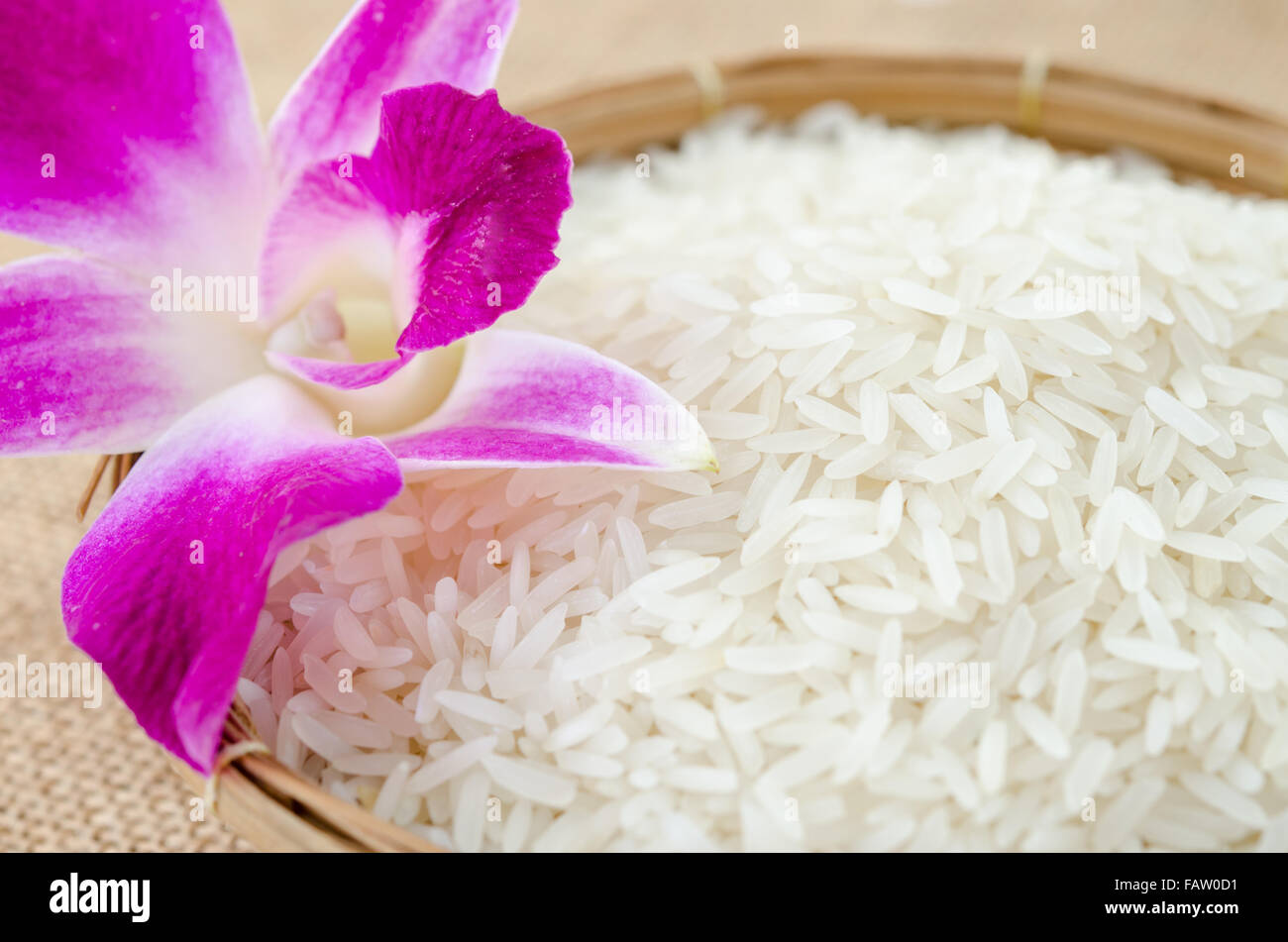 Raw rice with violet orchid flower in weave basket on sack background. Stock Photo