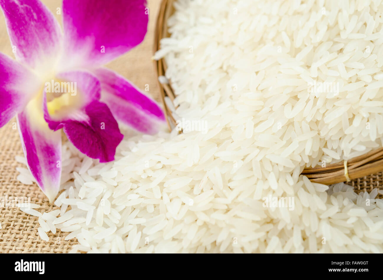 Raw rice with violet orchid flower in weave basket on sack background. Stock Photo