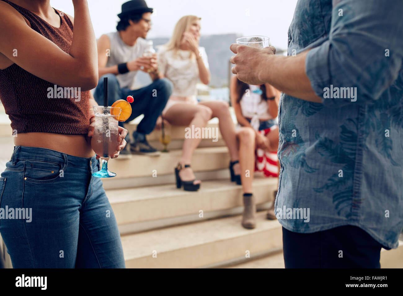 Cropped shot of a couple holding cocktails glasses while three people talking to each other in the background. Young people havi Stock Photo