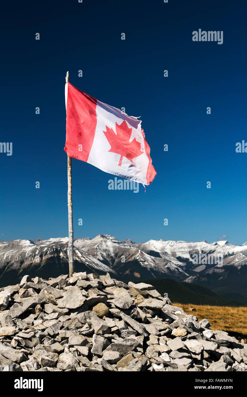 Well worn Canadian flag on wooden branch propped in a pile of rocks on top of a mountain with mountain range and blue sky in the background Stock Photo