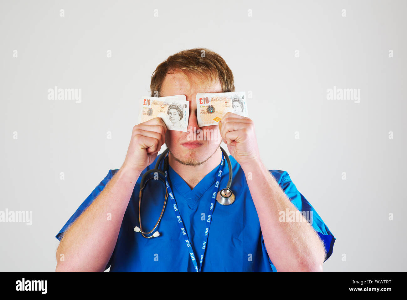 Young white male junior hospital doctor in theatre blues wearing a stethoscope holds a handful of ten pound notes to his face Stock Photo