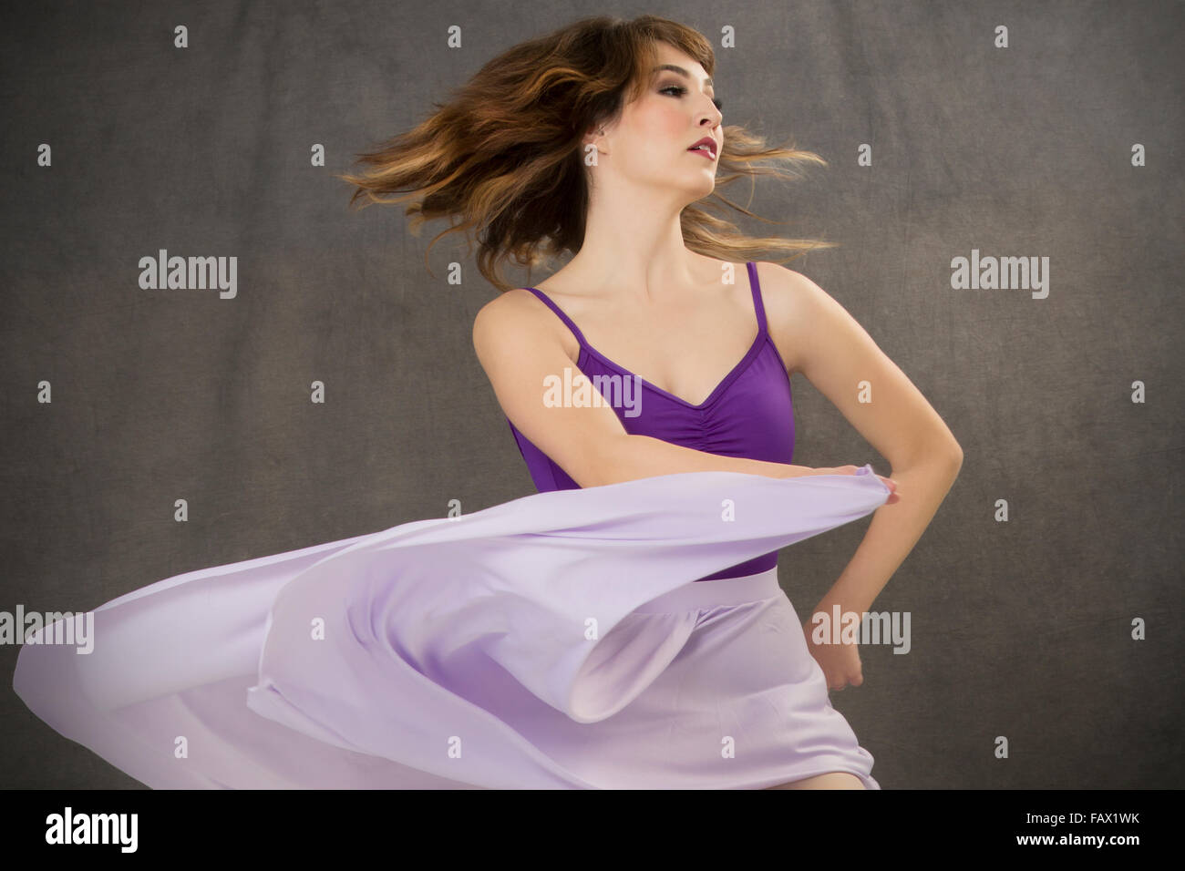 Attractive young woman dancer, turning in purple leotard and flowing lavender dress, from waist up on gray background. Stock Photo