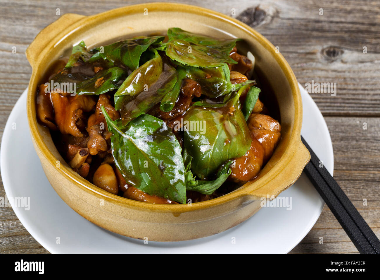 Close up front view of chicken and basil dish. Chopsticks on white plate underneath bowl on rustic wood background. Stock Photo