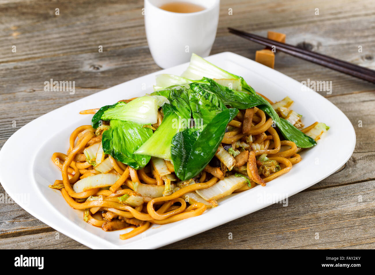 Close up front view of spicy cooked noodles, onion, bok choy, and chicken slices. Chopsticks and tea in background Stock Photo
