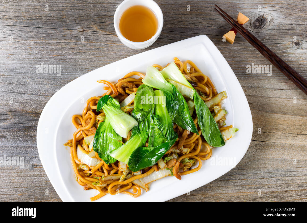 High angled view of spicy cooked noodles, onion, bok choy, chicken slices, chopsticks and tea on rustic wood. Stock Photo