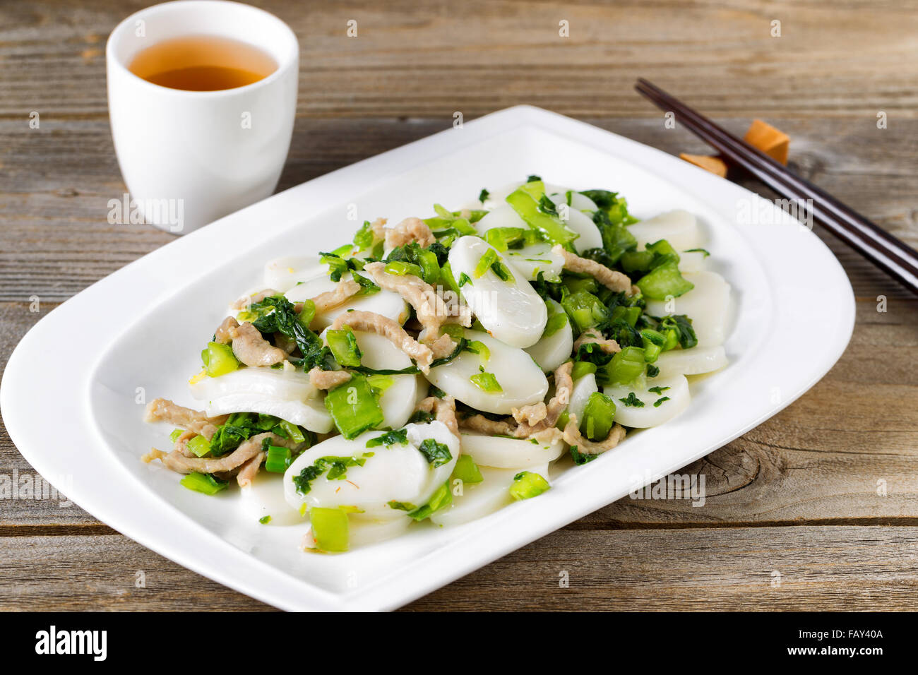 Close up front view of stir fry consisting of sliced sticky rice cakes, onion, and chicken. Chopsticks and tea in background on Stock Photo