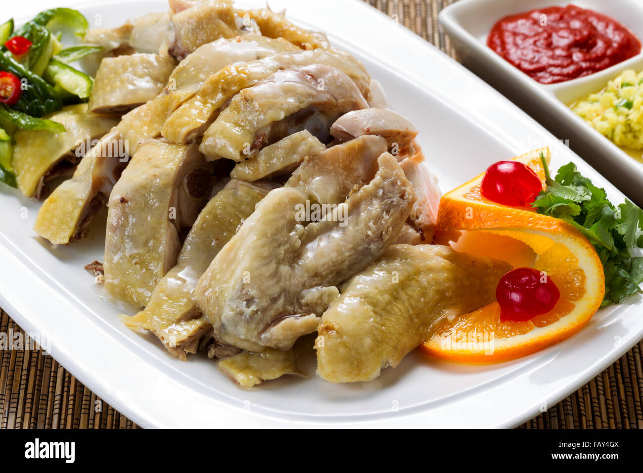 Close up front view of a succulent sliced chicken with dipping sauces and garnishes in background on bamboo mat. Stock Photo