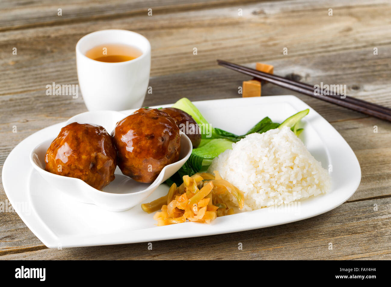 Close up front view Asian saucy meatballs, rice and bok choy on white plate. Chopsticks and green tea in background. Stock Photo