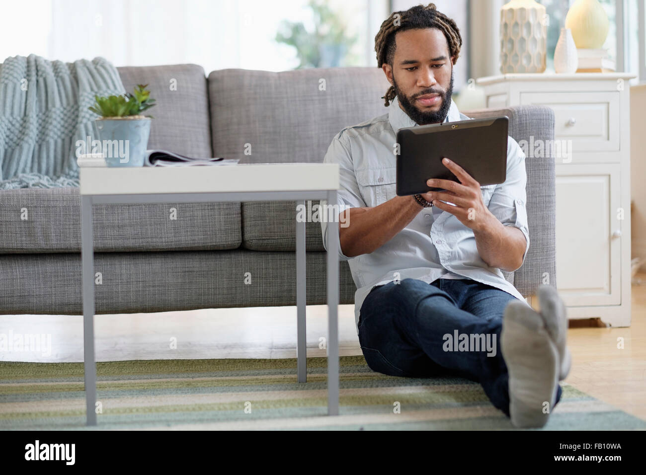 Young man with dreadlocks using digital table by sofa in living room Stock Photo