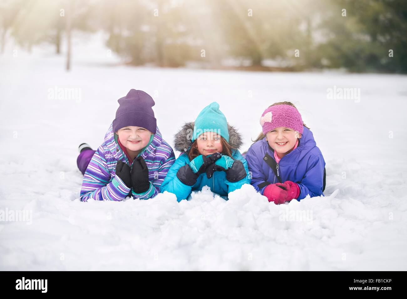 Friends wearing knit hats lying on front side by side in snow looking at camera smiling Stock Photo