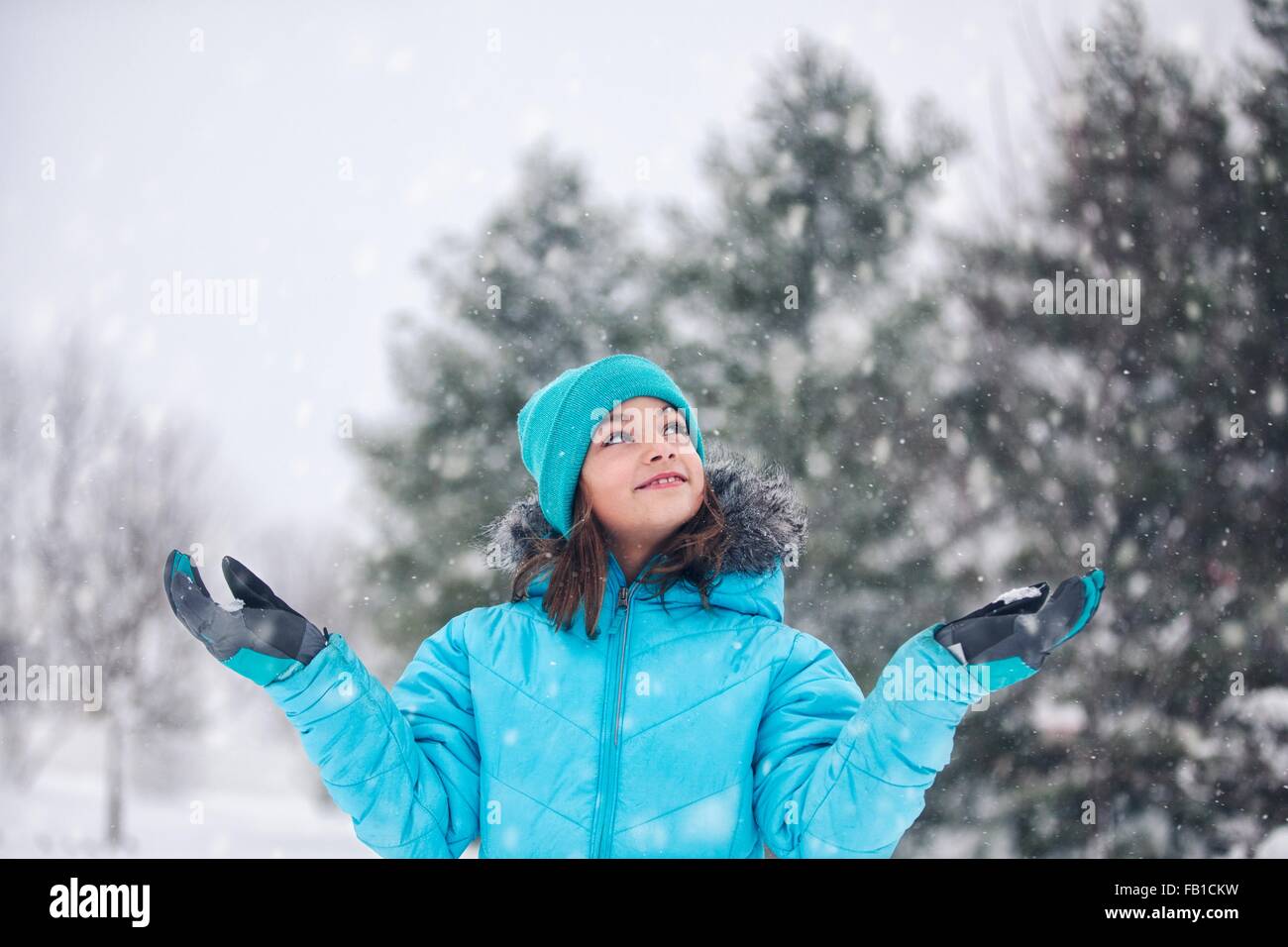 Girl wearing turquoise knit hat and coat, arms raised, hands out catching snow, looking up smiling Stock Photo