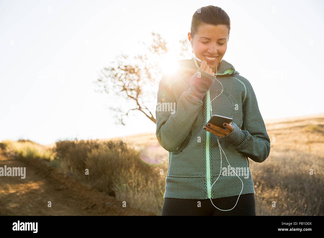Mature female runner on sunlit hill choosing smartphone music Stock Photo