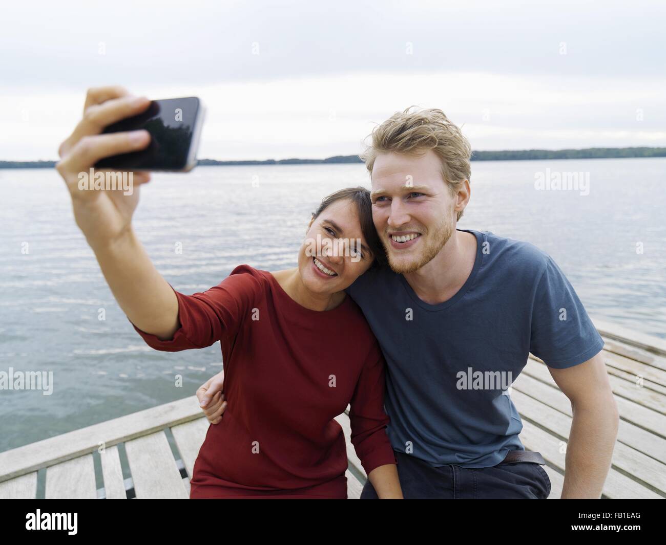 Couple on pier side by side using smartphone to take selfie smiling, Copenhagen, Denmark Stock Photo