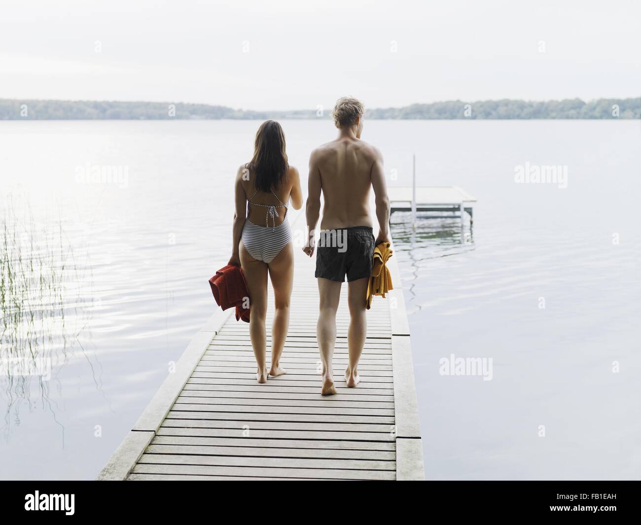 Rear view of couple wearing swimsuits, walking down pier carrying towels, Copenhagen, Denmark Stock Photo