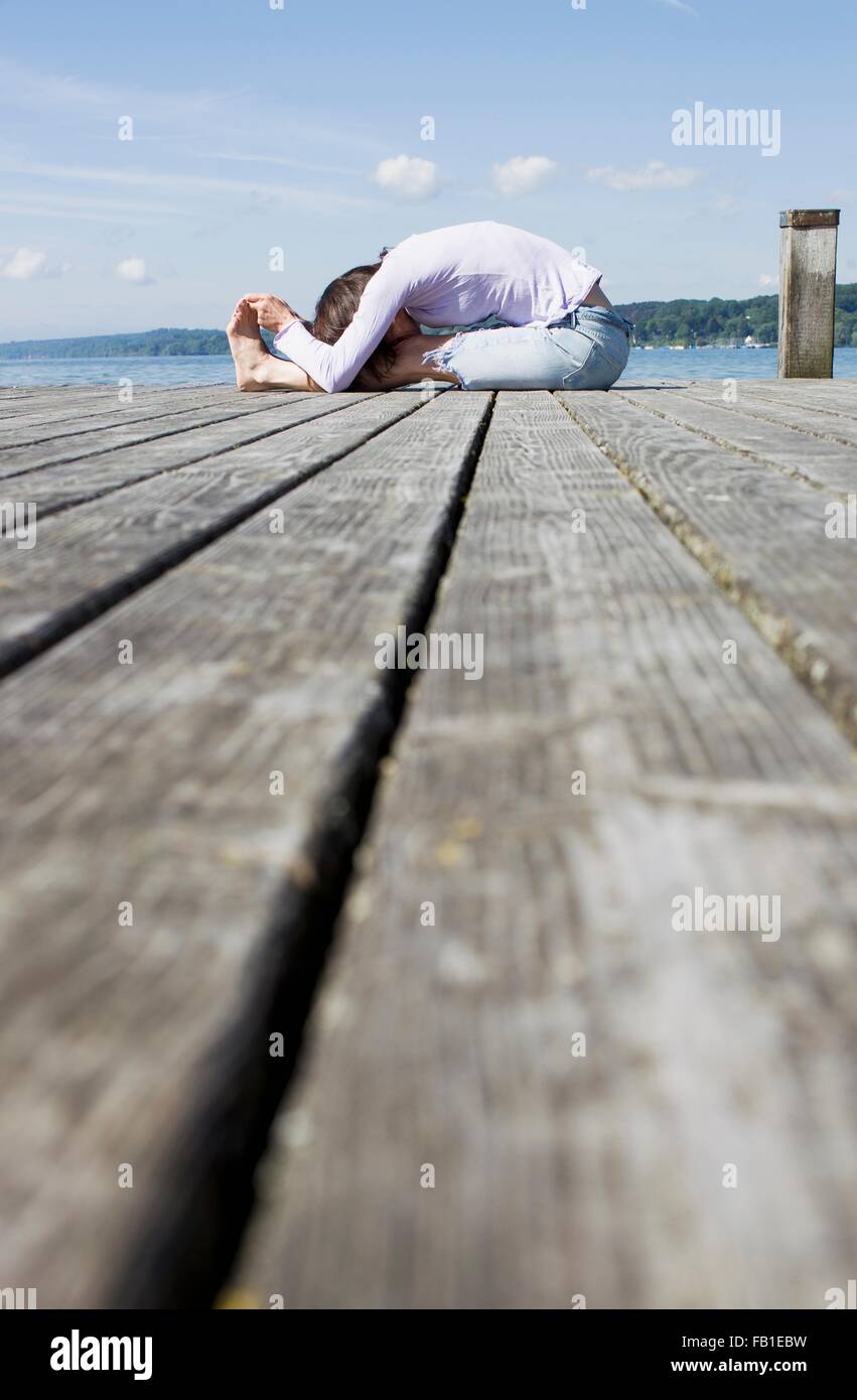 Surface level side view of mature woman sitting on pier bending over touching toes Stock Photo