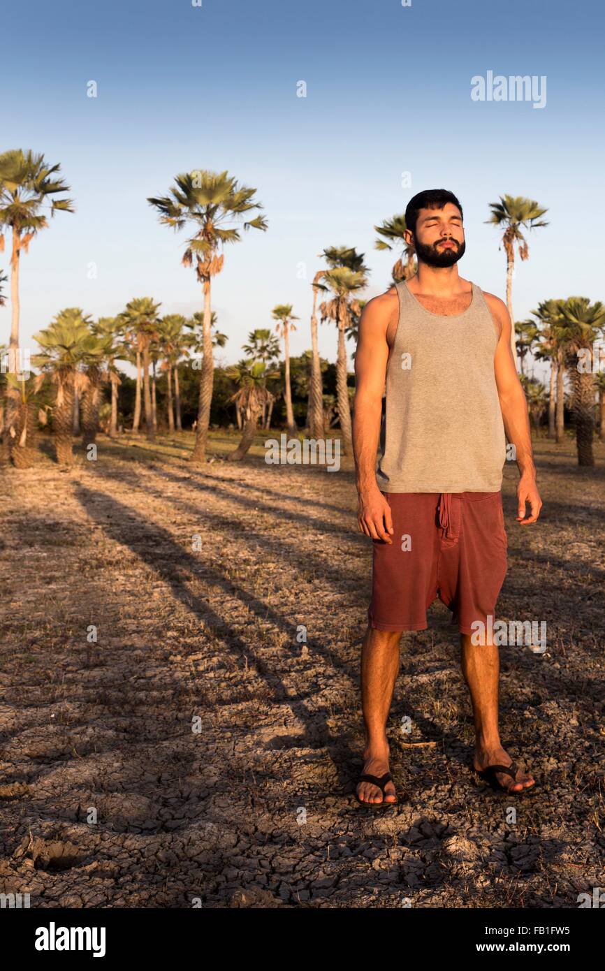 Full length front view of young man standing in front of palm trees casting shadow looking away, Taiba, Ceara, Brazil Stock Photo