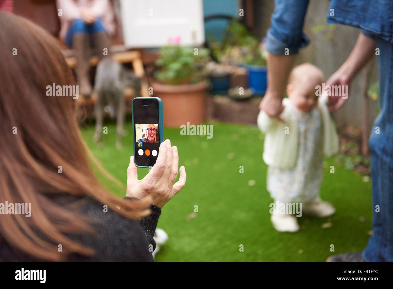 Mother in garden using smartphone to film daughter learning to walk, focus on foreground Stock Photo