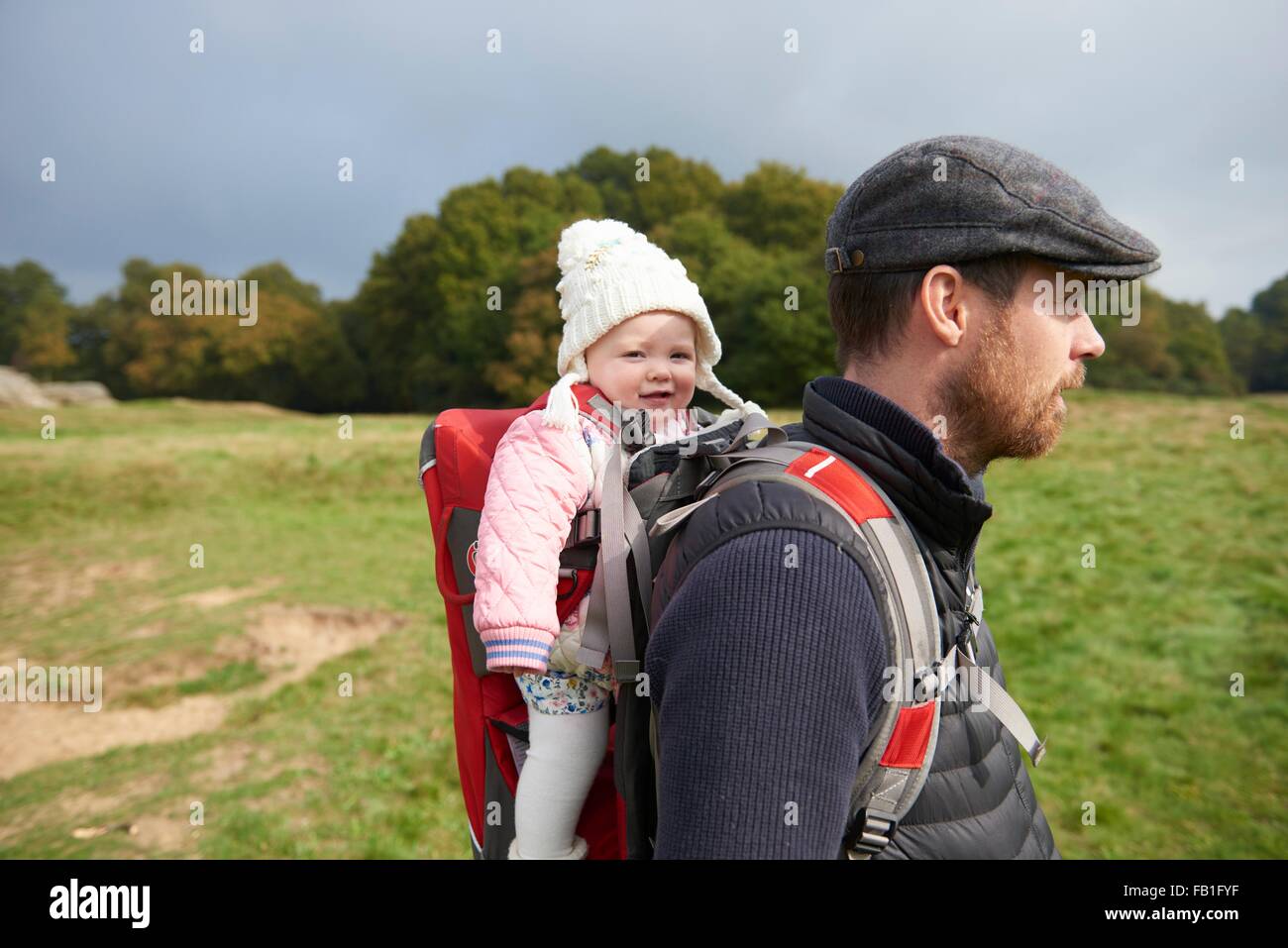 Side view of mid adult man in field wearing flat cap carrying daughter on back in baby carrier Stock Photo
