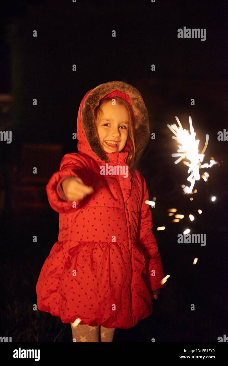 Girl wearing red fur trim coat holding sparkler, looking at camera smiling Stock Photo