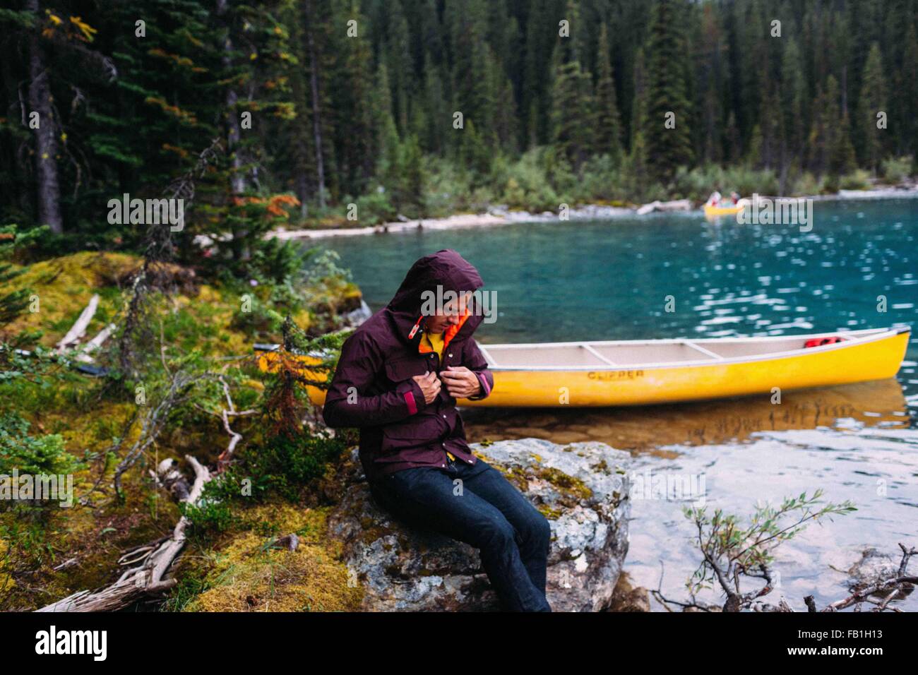 Mid adult man sitting by canoe on Moraine lake, looking down fastening waterproof coat, Banff National Park, Alberta Canada Stock Photo