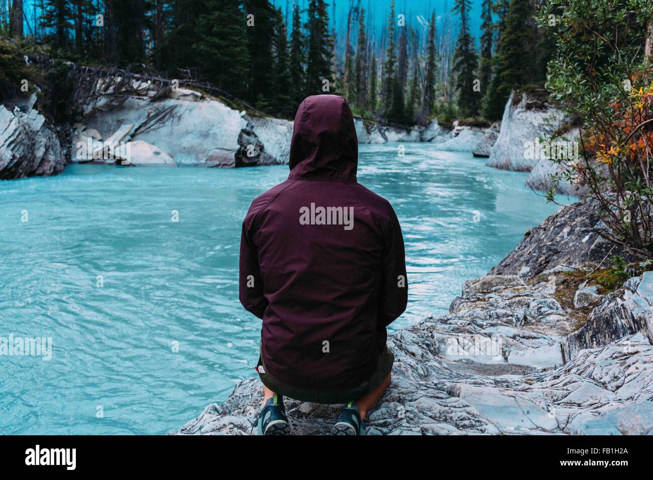 Rear view mid adult man wearing hooded waterproof coat crouching waters edge Moraine lake Banff National Park Alberta Canada Stock Photo