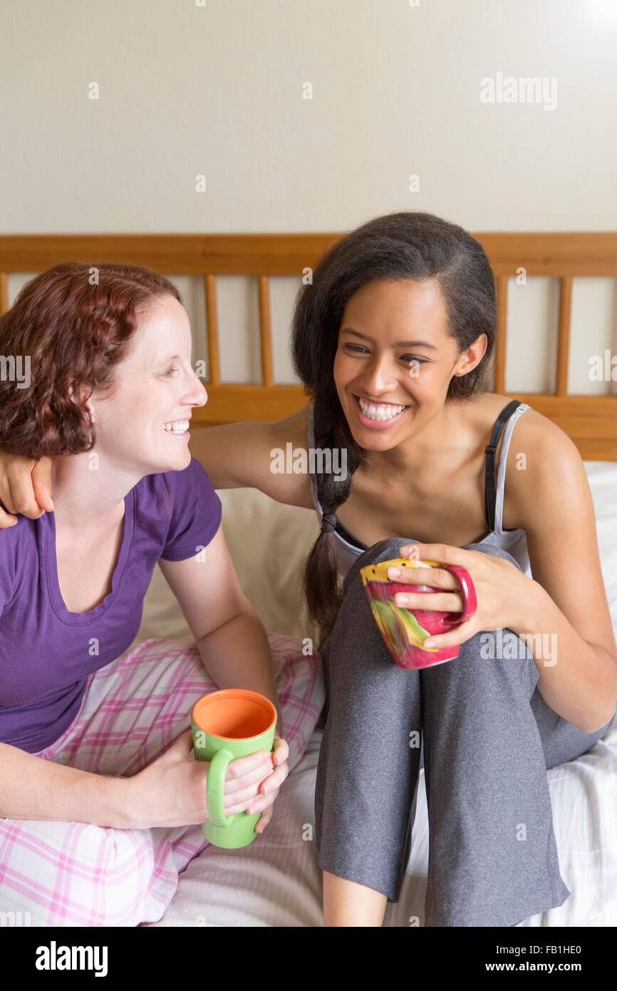 Young women sitting on bed, arm around friend, holding cup smiling Stock Photo
