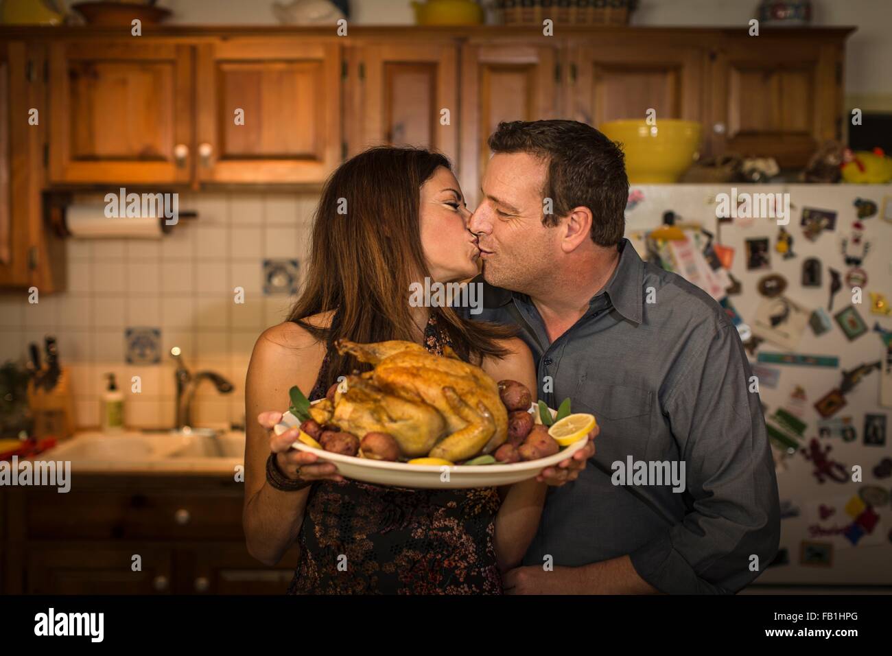 Mature couple kissing whilst holding roast chicken dinner in kitchen Stock Photo
