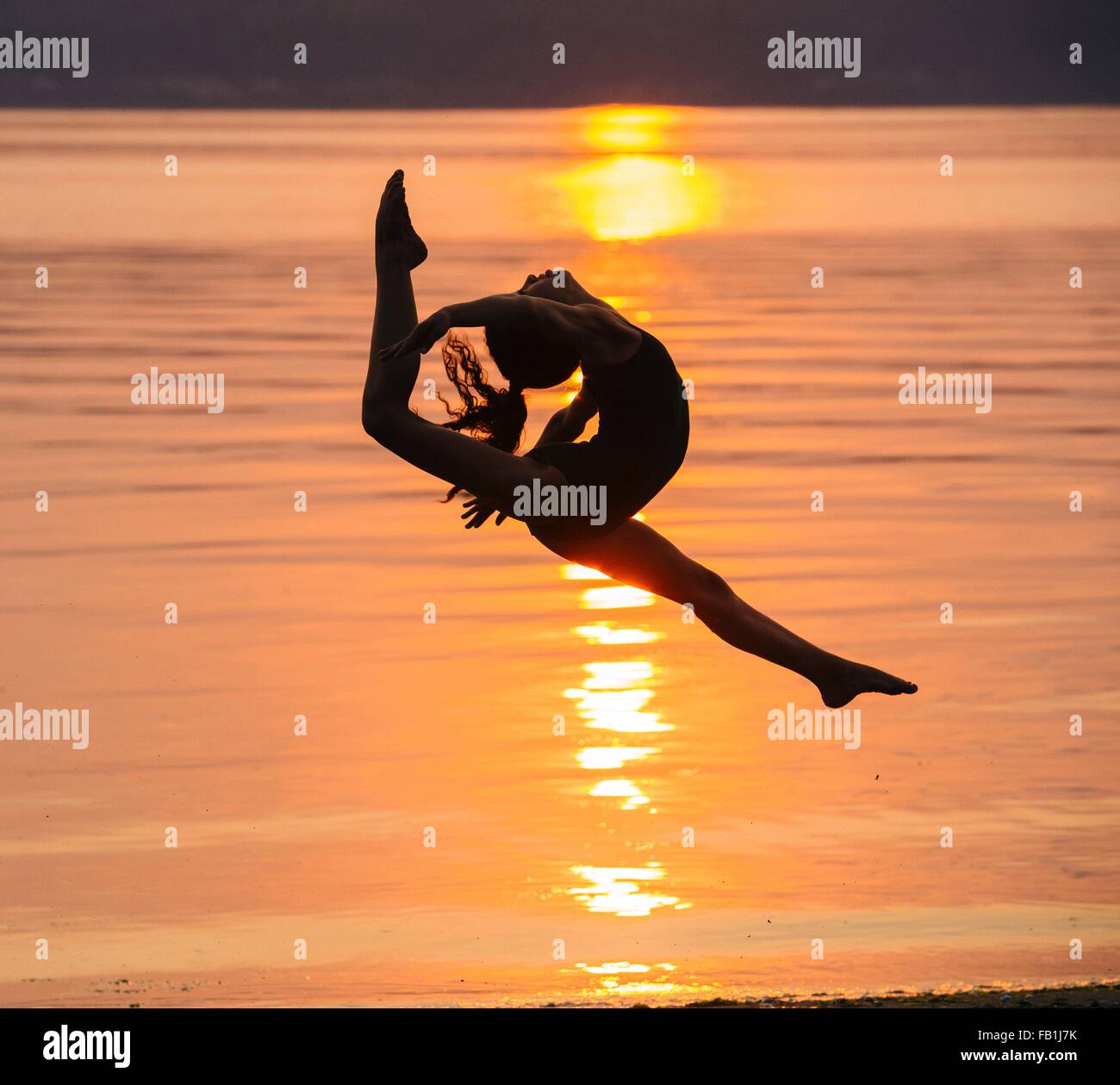Side view of girl in silhouette by ocean at sunset in mid air, legs apart throwing head back Stock Photo