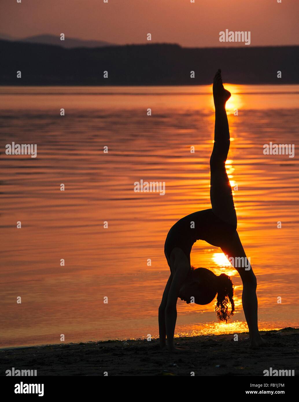 Side view of girl in silhouette by ocean at sunset bending over backwards leg raised doing the splits Stock Photo