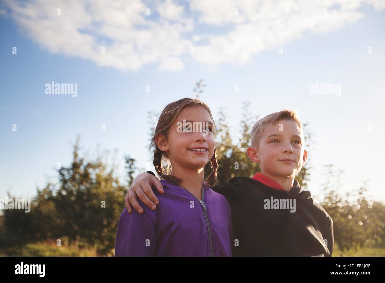 Low angle view of boy outdoors with arms around girl looking away smiling Stock Photo