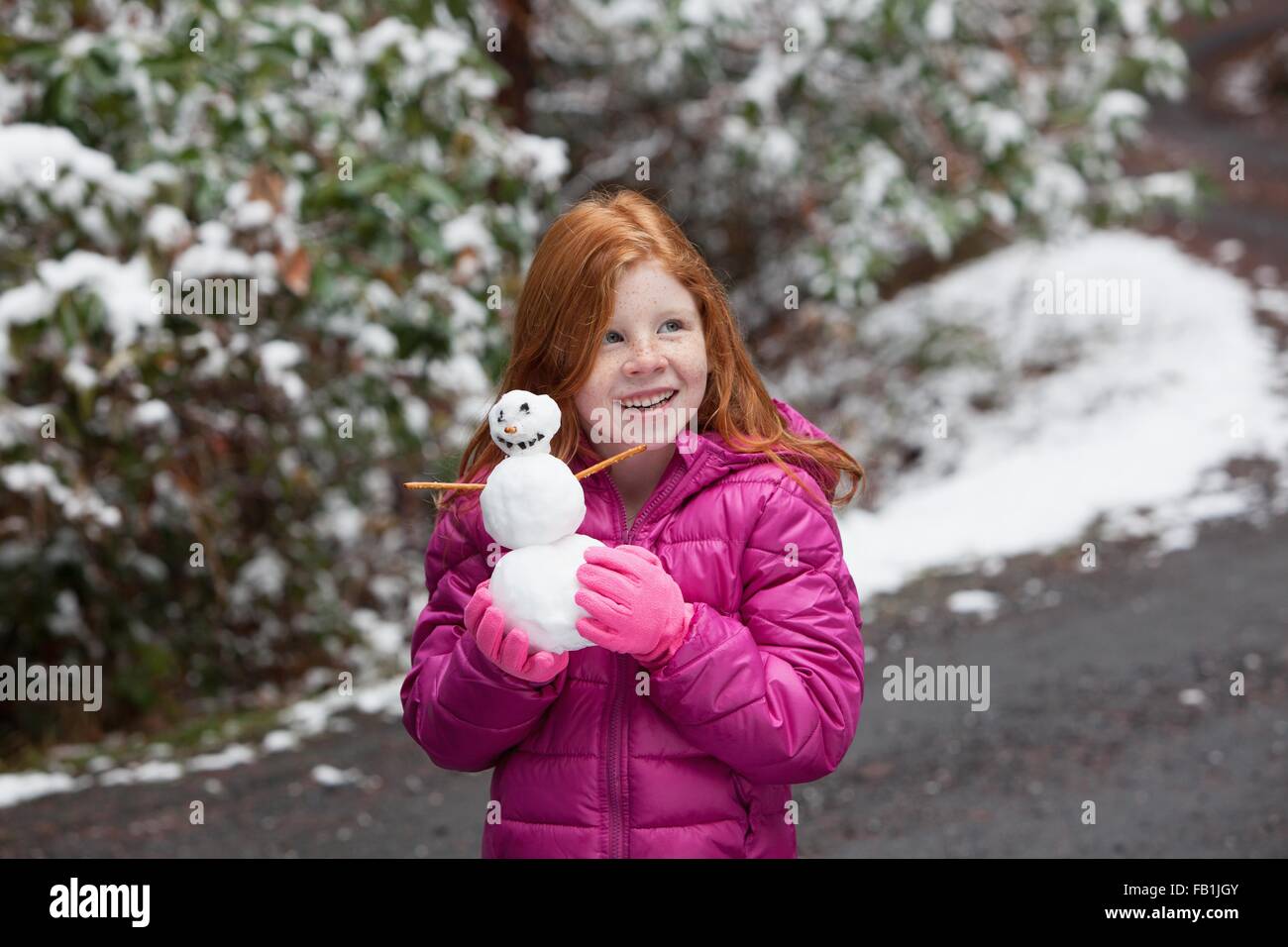 Red haired girl in front of snow covered trees, wearing pink gloves and padded jacket holding snowman looking away smiling Stock Photo