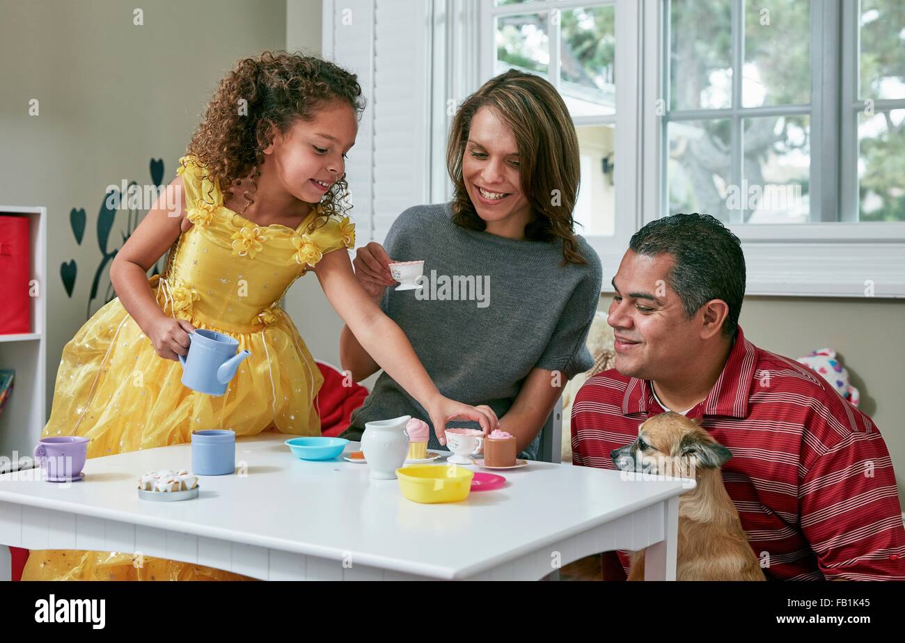 Girl in playroom sitting at table serving tea from toy tea set to parents Stock Photo