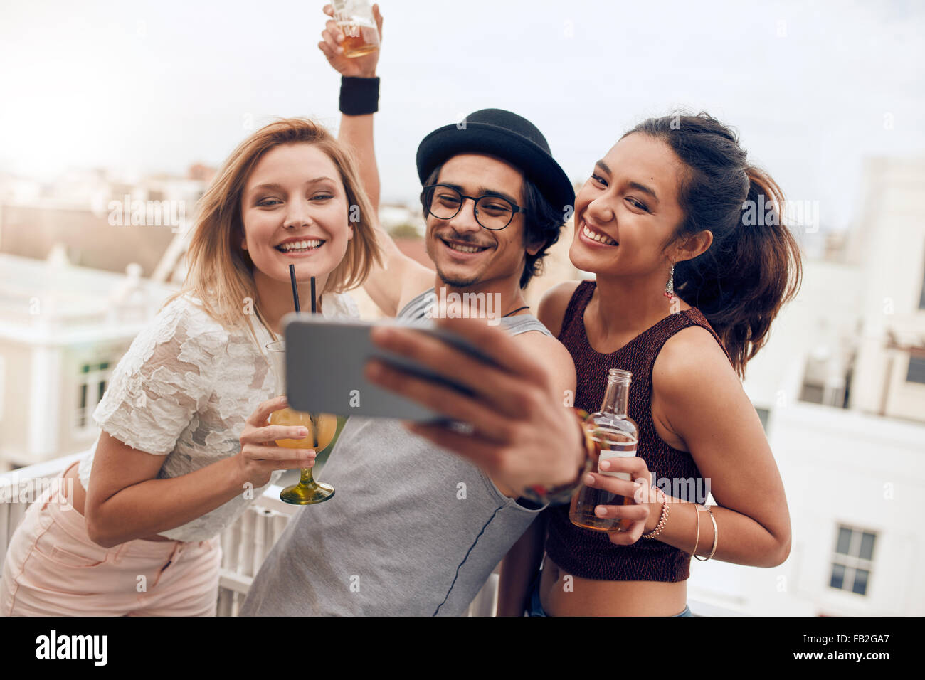 Three young friends taking self portrait with smart phone during a party. Happy young man and woman taking selfie at rooftop par Stock Photo
