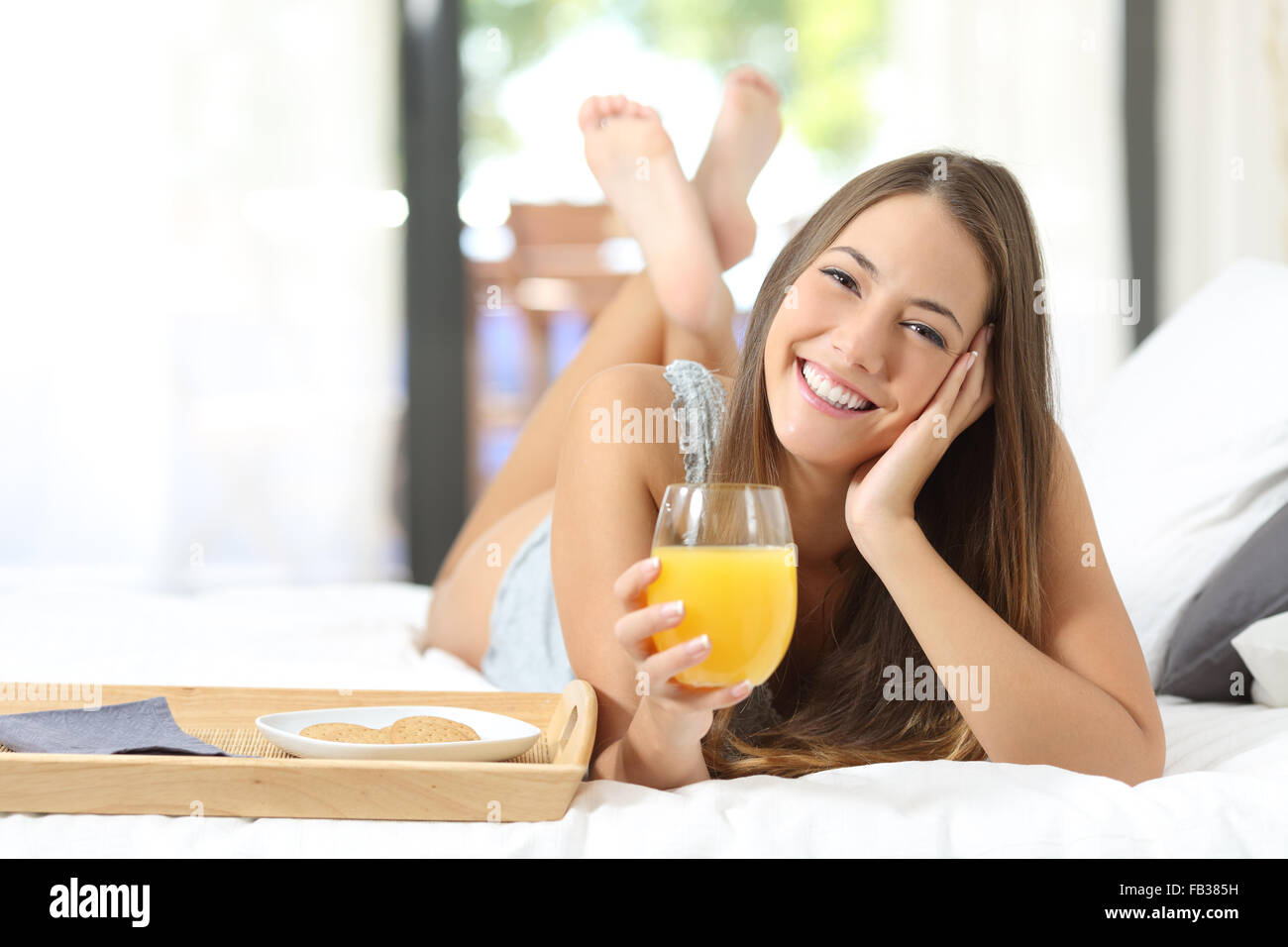 Happy girl having breakfast with orange juice on the bed at home and looking at camera Stock Photo