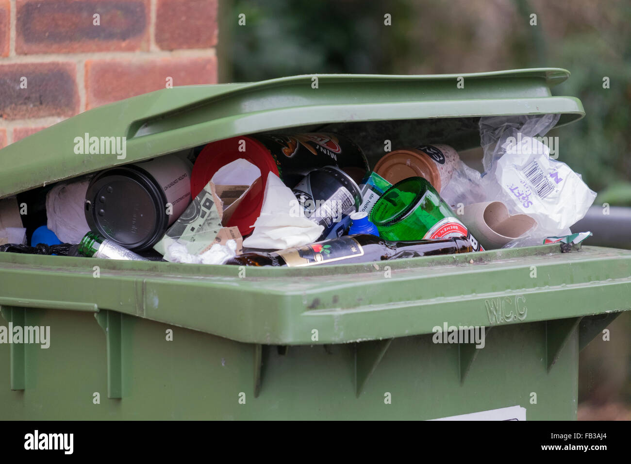 green refuse wheelie bin overflowing with glass bottles tins and paper